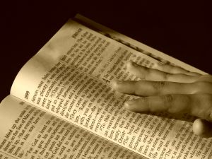 sepia tones, bible in forground and left, right hand holding it stretched across the right handed page